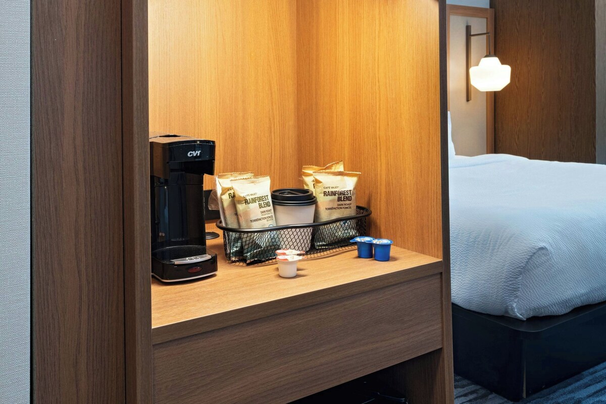 Habitación de hotel con un microondas en un estante de madera, una cafetera y una bandeja con cápsulas de café y tazas.