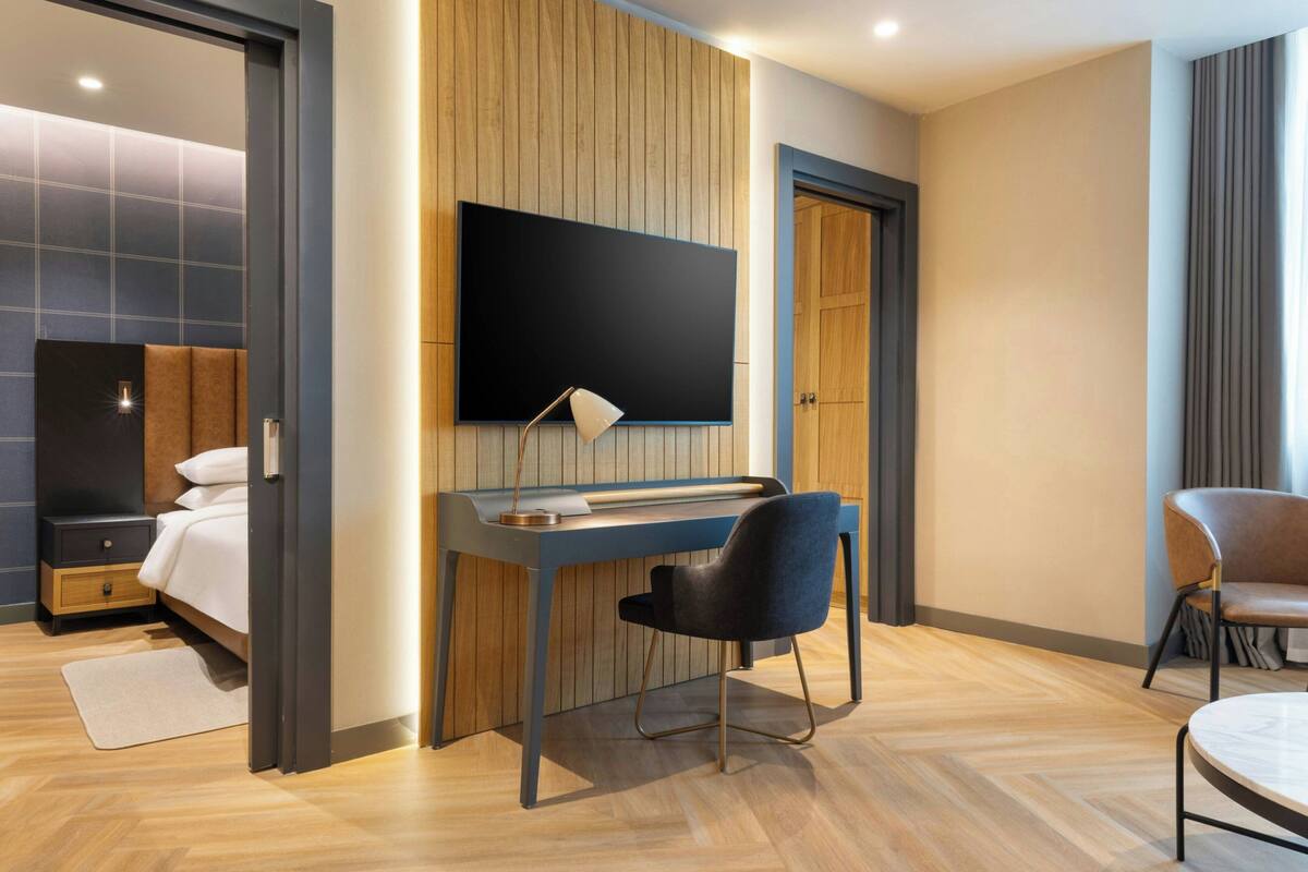 Ett modernt hotellrum med en säng, ett skrivbord med en stol, en tv som är monterad på väggen och en dörr.