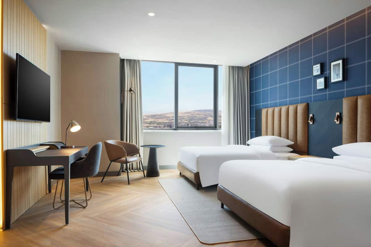 Ett hotellrum med en stor säng, ett skrivbord med en stol, en TV och utsikt över staden.