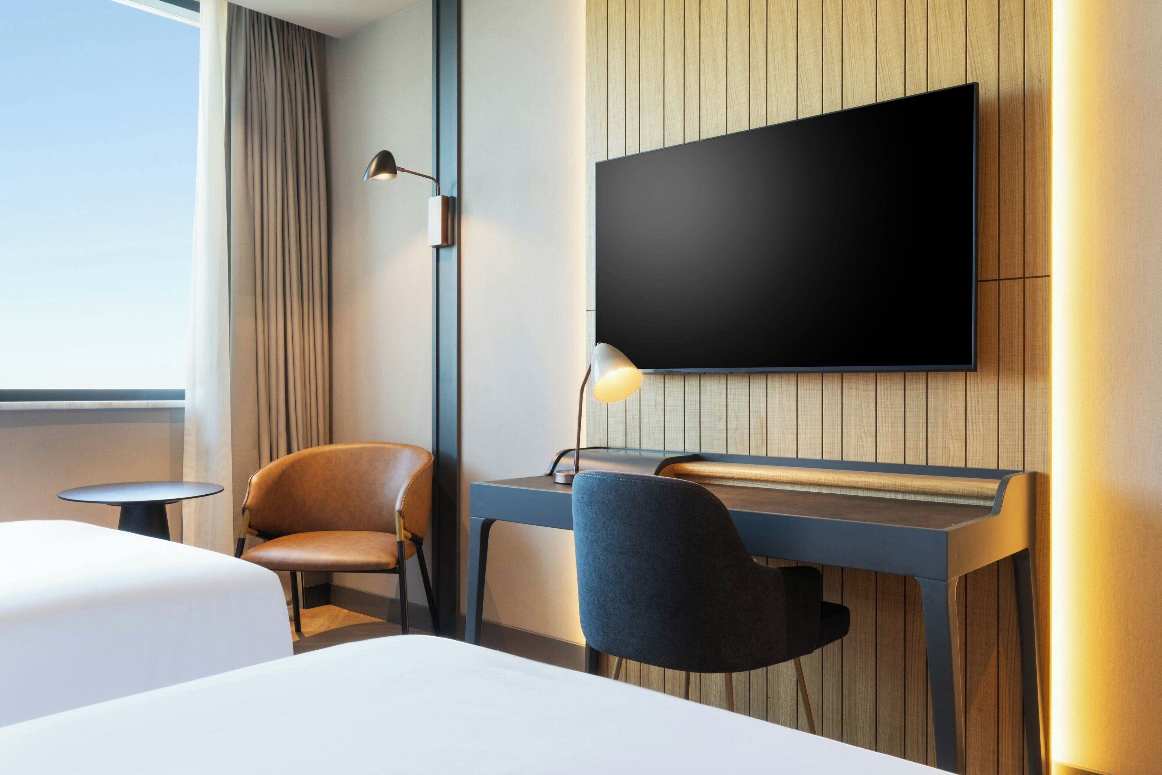 A hotel room with a large flat-screen TV mounted on the wall, a desk with a chair, a bed with white bedding, and a small round table.
