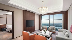 Una sala de estar moderna con un televisor grande, asientos cómodos, una araña y vistas de un paisaje urbano a través de grandes ventanales.