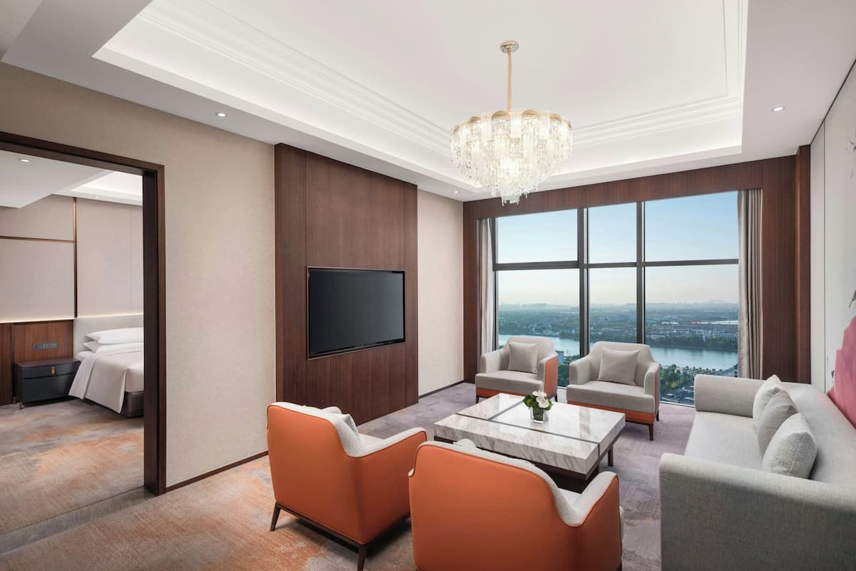 A modern living room with a large TV, comfortable seating, a chandelier, and a view of a cityscape through large windows.