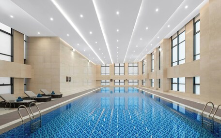 A large indoor swimming pool with clear blue water, surrounded by lounge chairs and sunbeds, with large windows and a modern ceiling design.