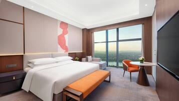 Una habitación de hotel moderna con una cama grande, un banco, una zona de estar con una mesa y un ventanal con vistas a la ciudad.
