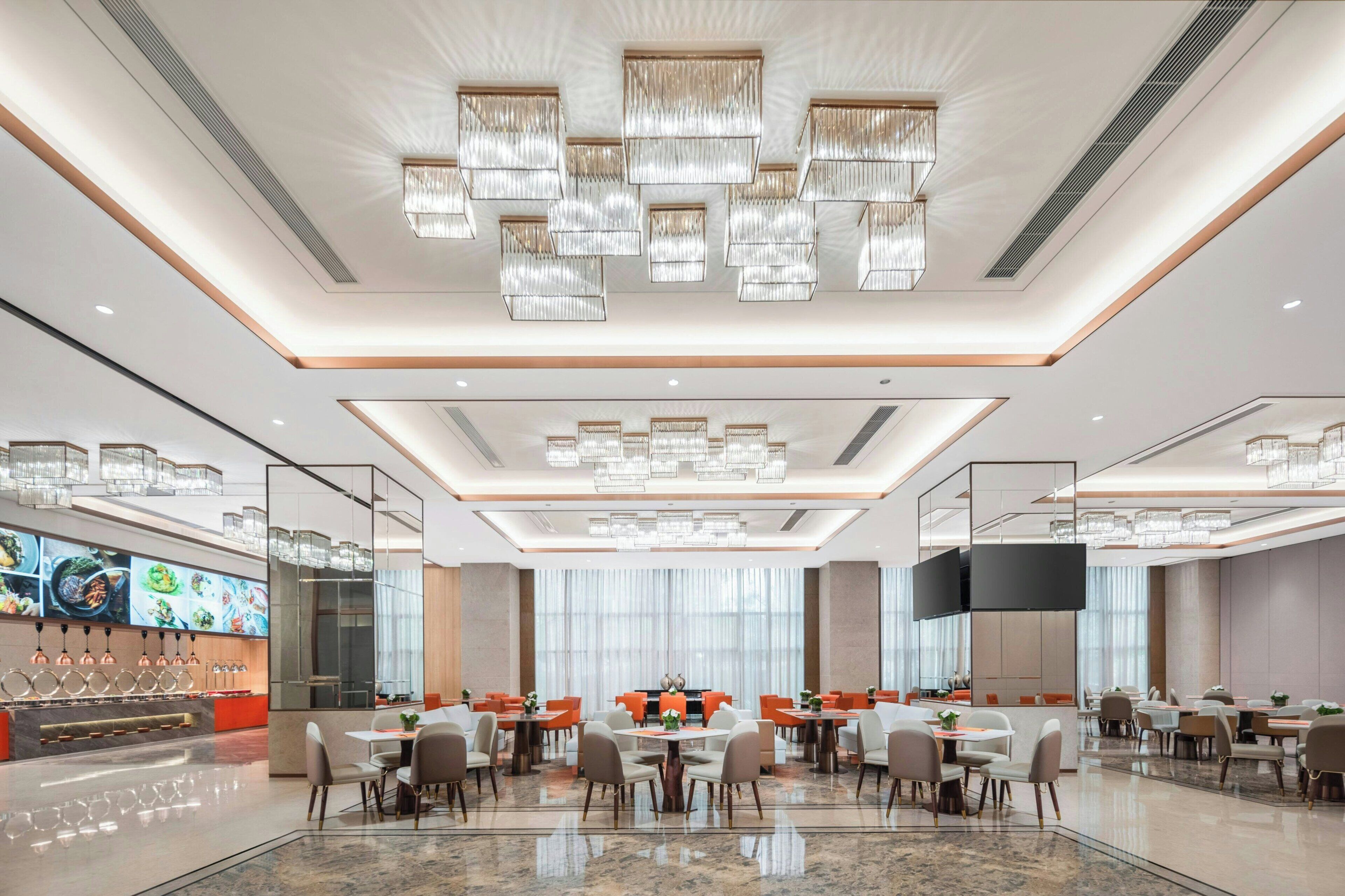 En modern hotellrestaurang med flera bord och stolar, en stor kristallkrona och en tv-skärm.