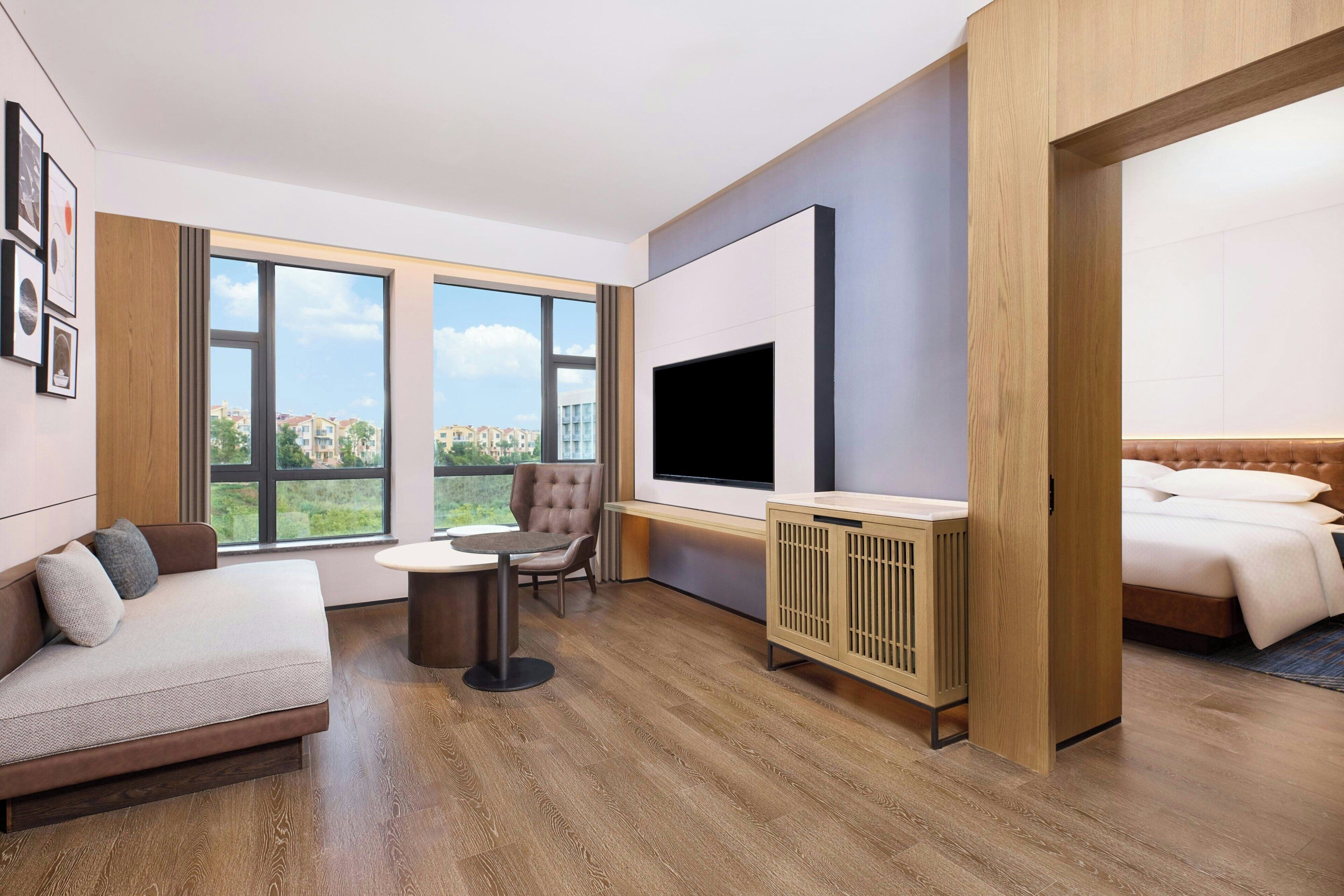 A modern hotel room with a sofa, a chair, a TV, a bed, and large windows offering a city view.