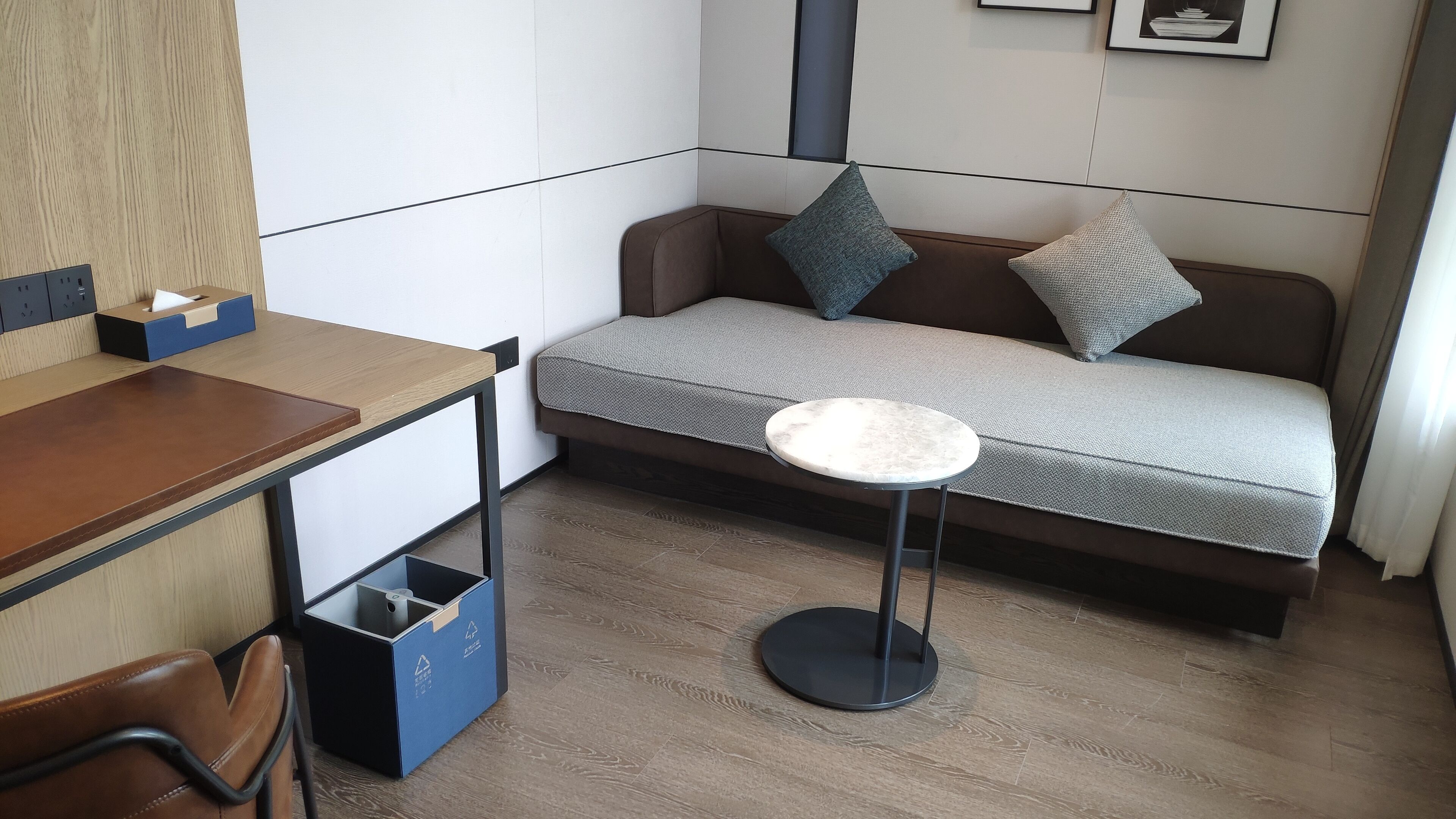 A compact room with a wooden desk, a small round table, a sofa with cushions, and a trash bin.