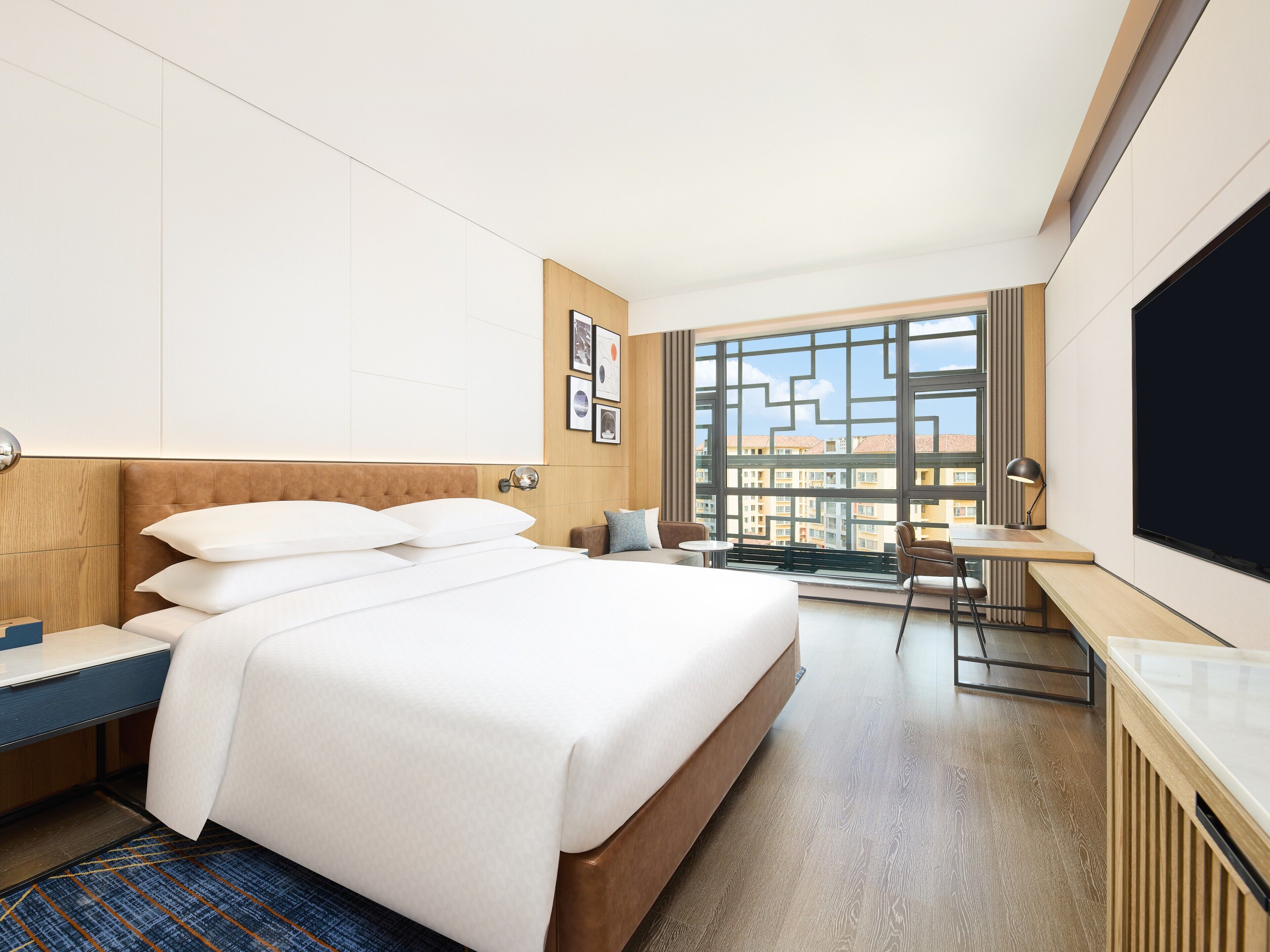 Moderni hotellihuone, jossa on suuri sänky, työpöytä ja suuri ikkuna, josta avautuu näkymä rakennuksiin.