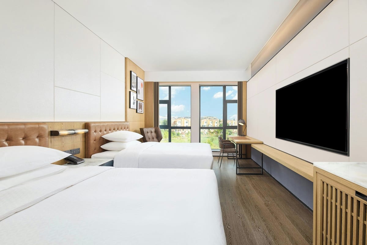 A hotel room with two beds, a desk, a chair, a TV, and large windows.