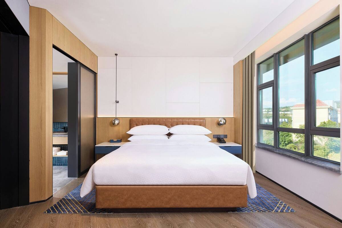 A modern hotel room with a large bed, wooden headboard, and a view through floor-to-ceiling windows.