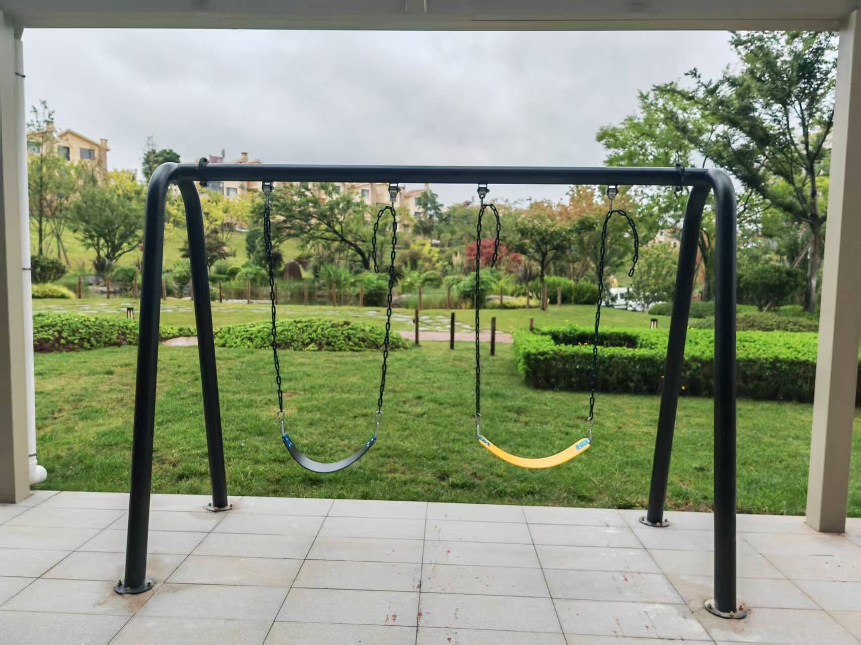 Un columpio en un parque con suelo de baldosas y follaje al fondo.