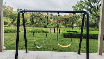 Un columpio en un parque con suelo de baldosas y follaje al fondo.