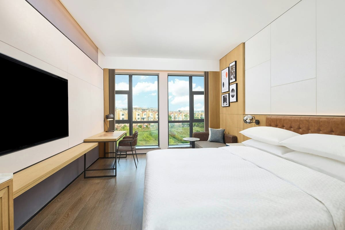 A hotel room with a large bed, a desk, and a TV. There are large windows with a view of buildings and greenery.