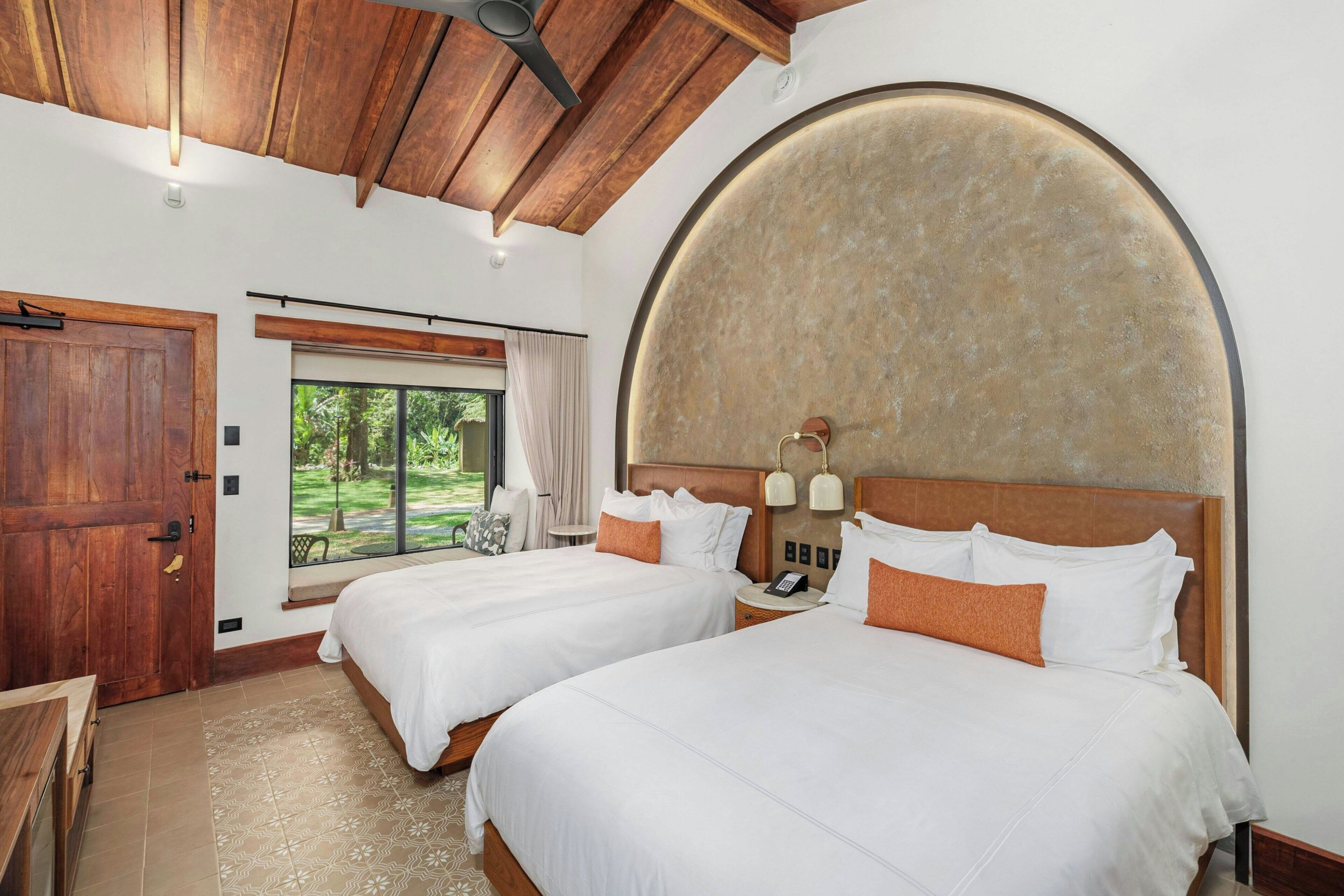 A hotel room with two beds, a wooden ceiling, a large window, and a round wall feature.
