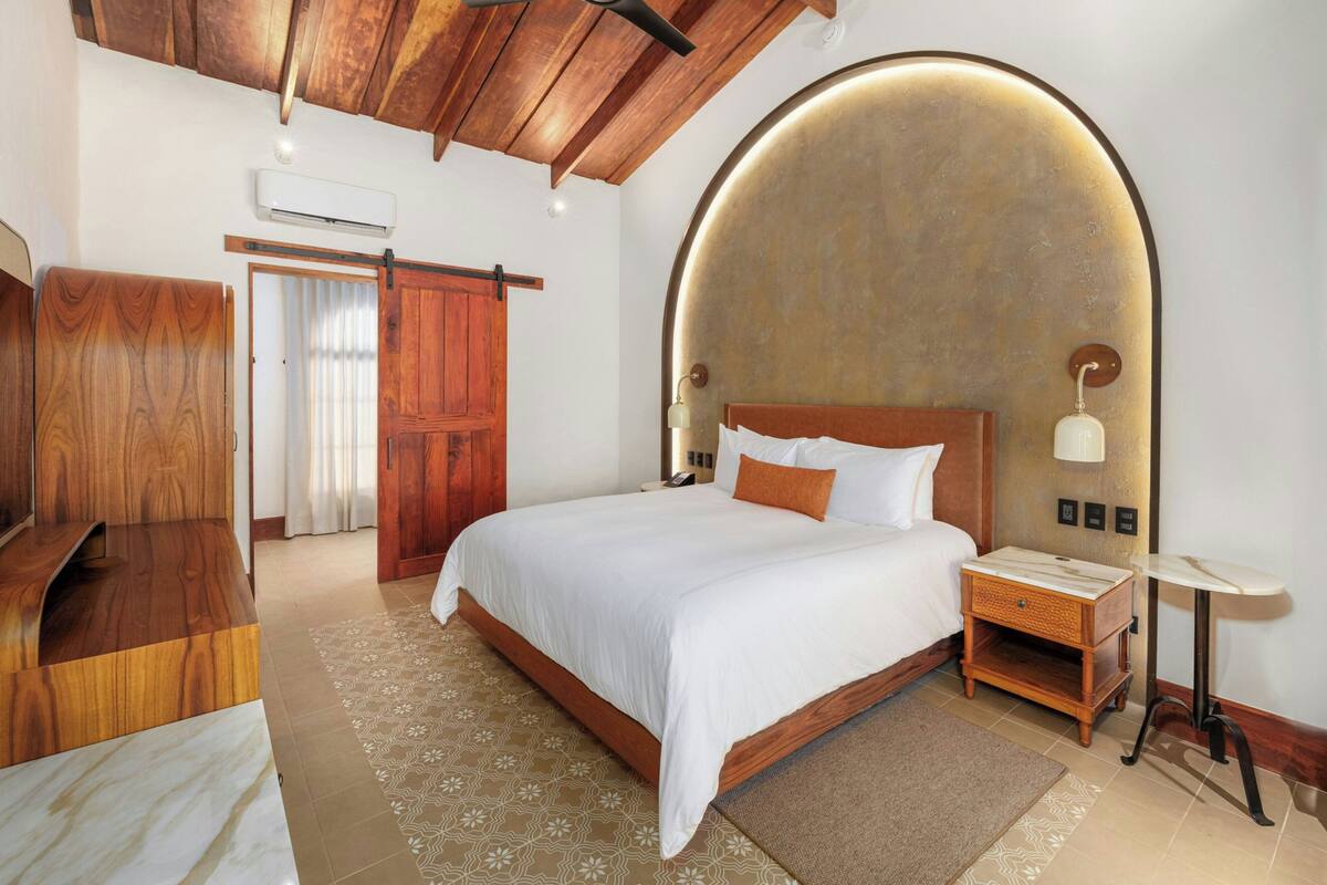 Quarto com uma cama grande, cabeceira de madeira, mesa de cabeceira e luminária fixada na parede.