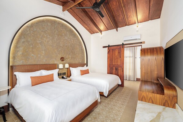 Habitación de hotel con dos camas, un llamativo elemento de pared curvo, ventilador de techo, televisor y armario de madera.