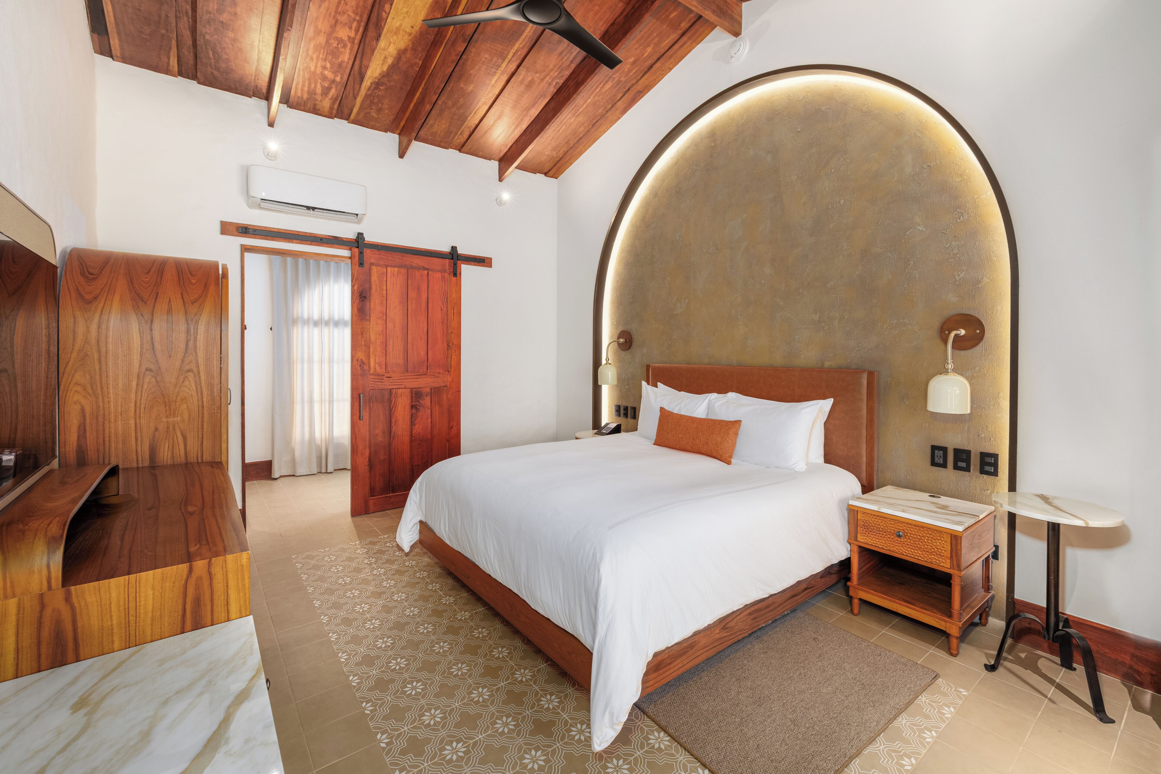 A bedroom with a wooden ceiling, a bed with white bedding, a nightstand, a wall-mounted lamp, and a wooden wardrobe.