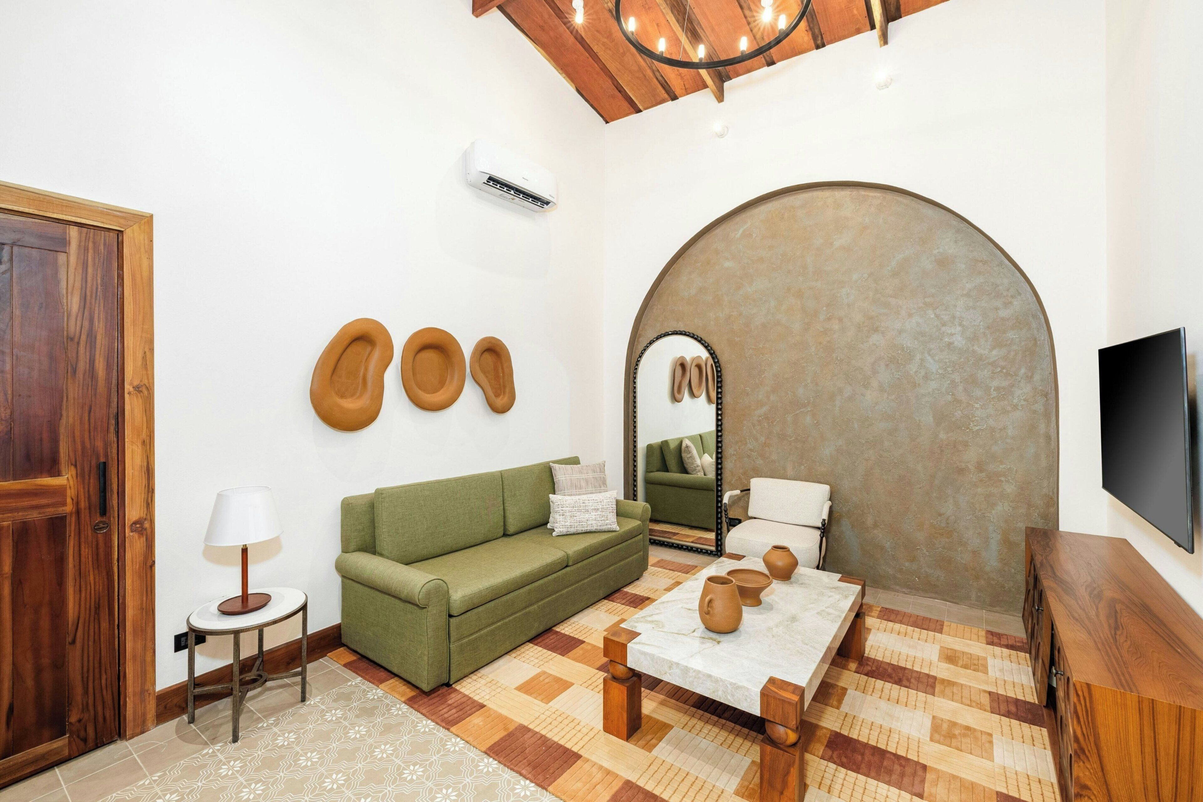 A room with a wooden door, a green sofa, a small table with pottery, a mirror, and a TV on the wall.