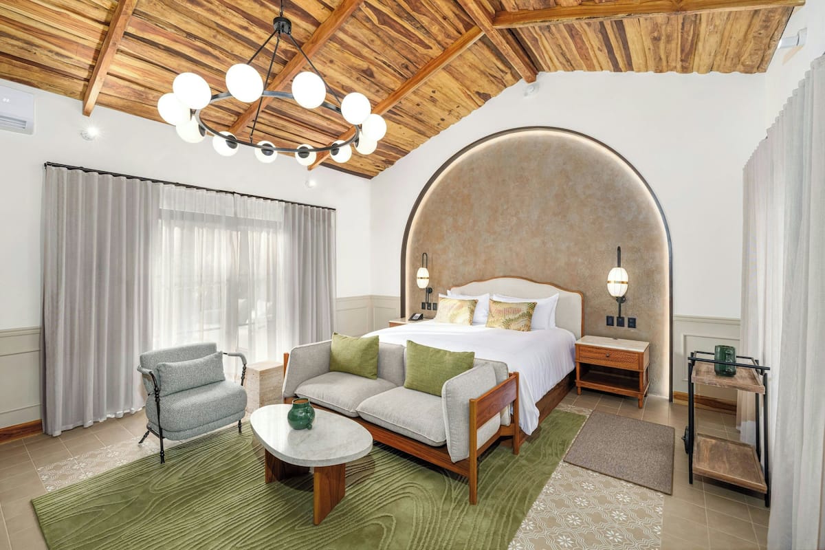 Quarto de hotel com cama, sofá, mesa pequena, mesa de cabeceira e ventilador de teto.