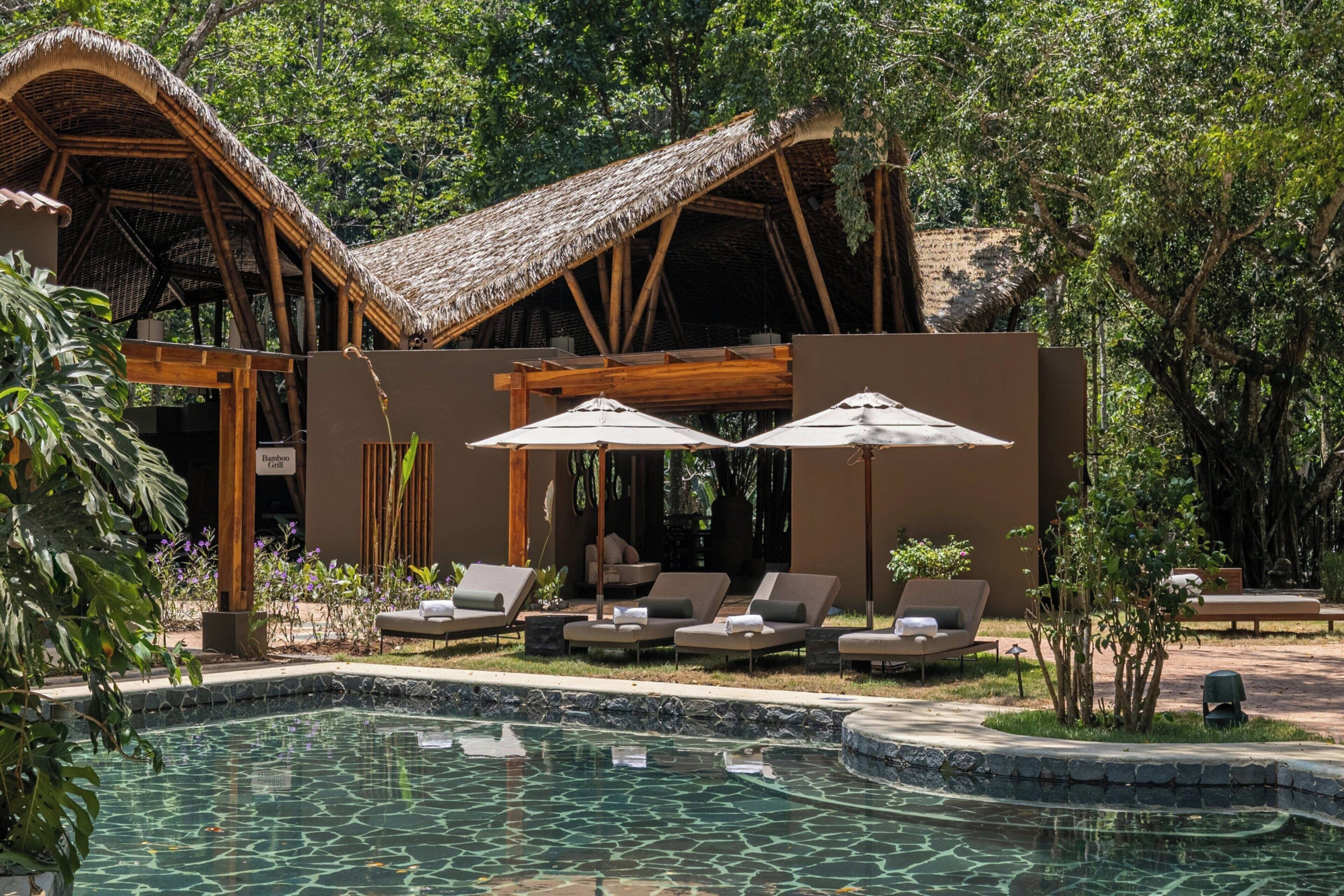 A pool area with lounge chairs and umbrellas, a bamboo grill, and a thatched-roof structure.