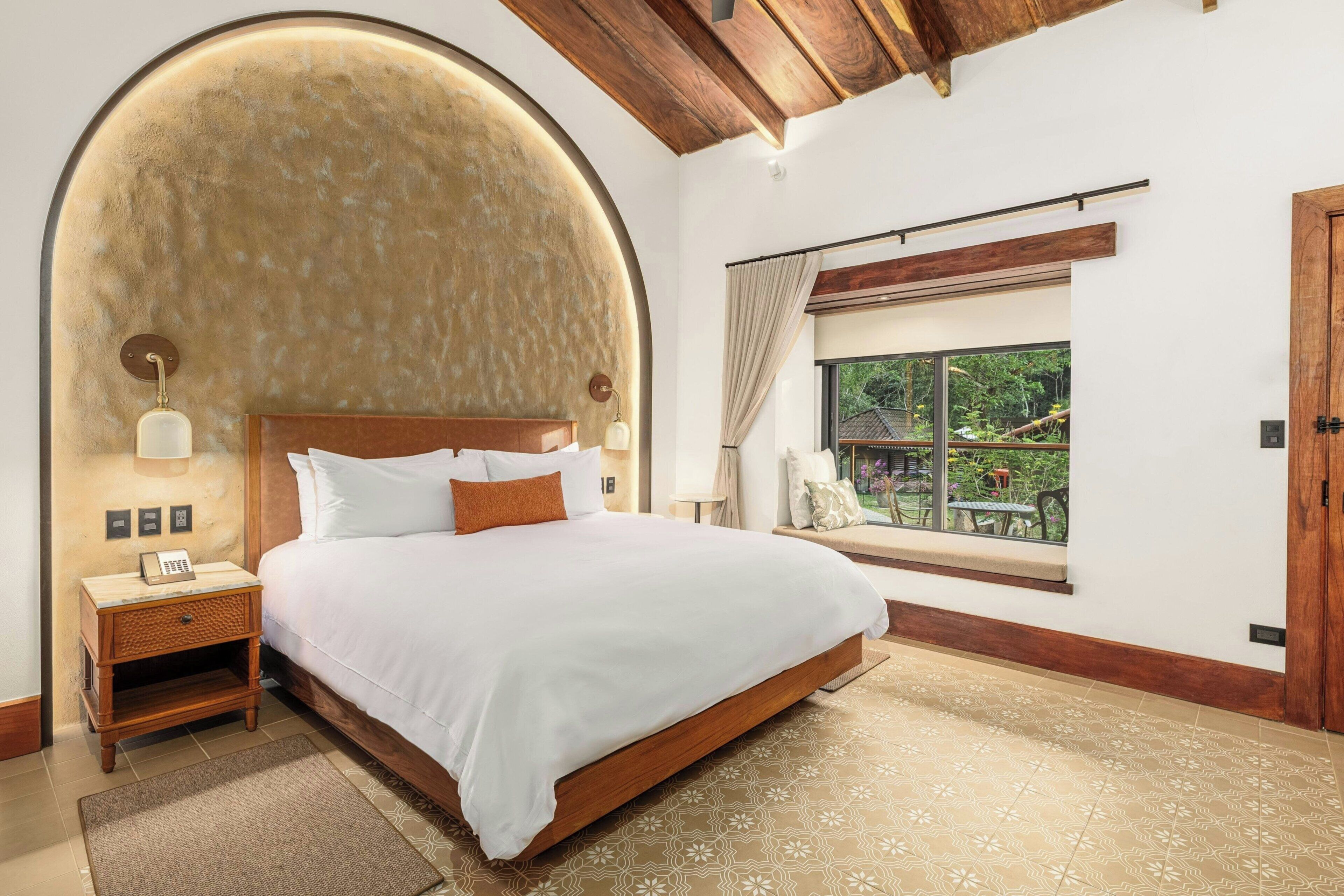 Quarto com cama grande, mesa de cabeceira, luminária, janela com vista e teto de madeira.