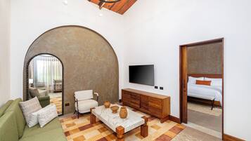 Una sala de estar moderna con sofá, mesa de centro, televisor y un amplio arco que da a un dormitorio con cama.