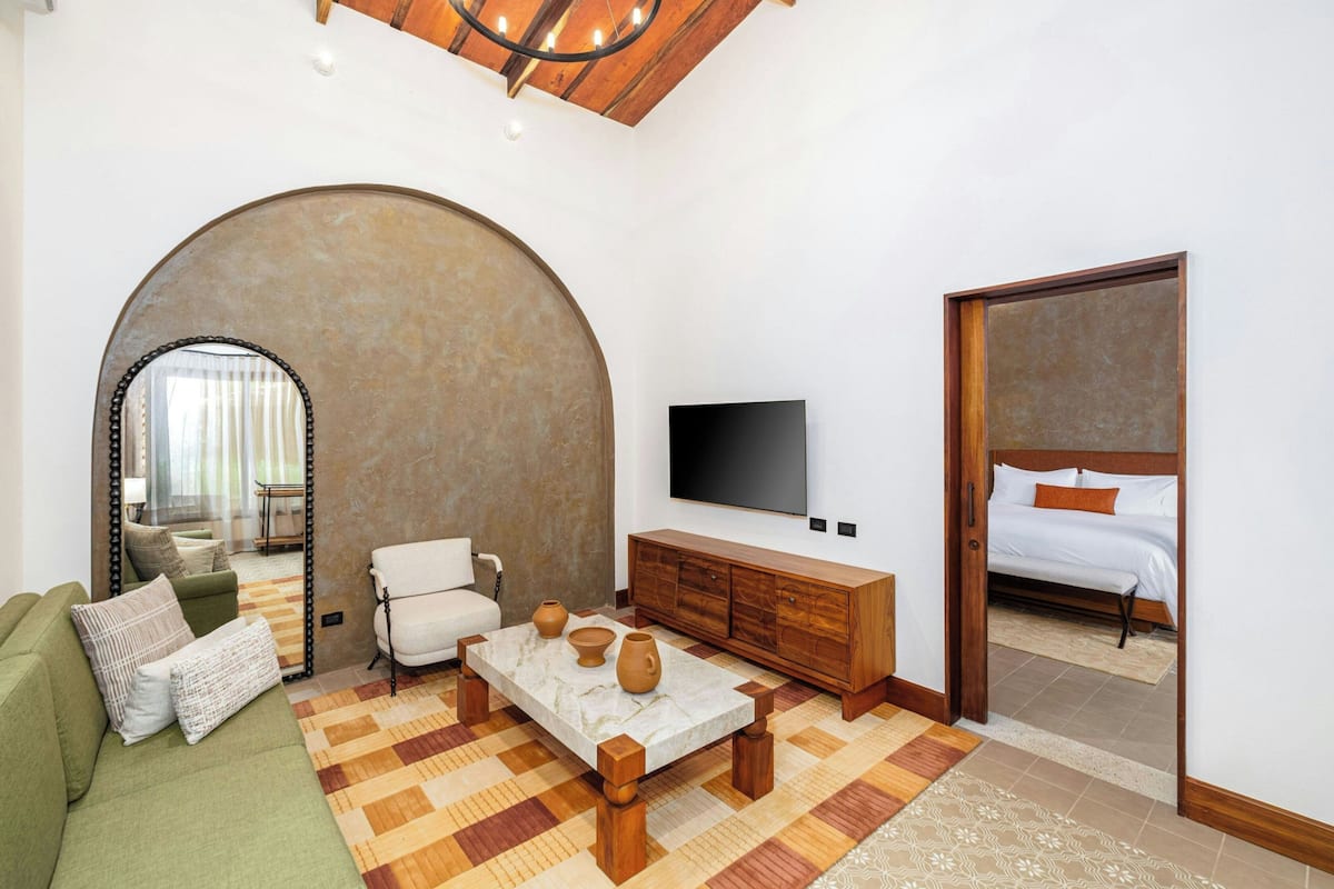 Quarto de hotel moderno com um sofá verde, uma mesa de centro de madeira, uma televisão e uma grande porta em arco que dá para um quarto com uma cama.