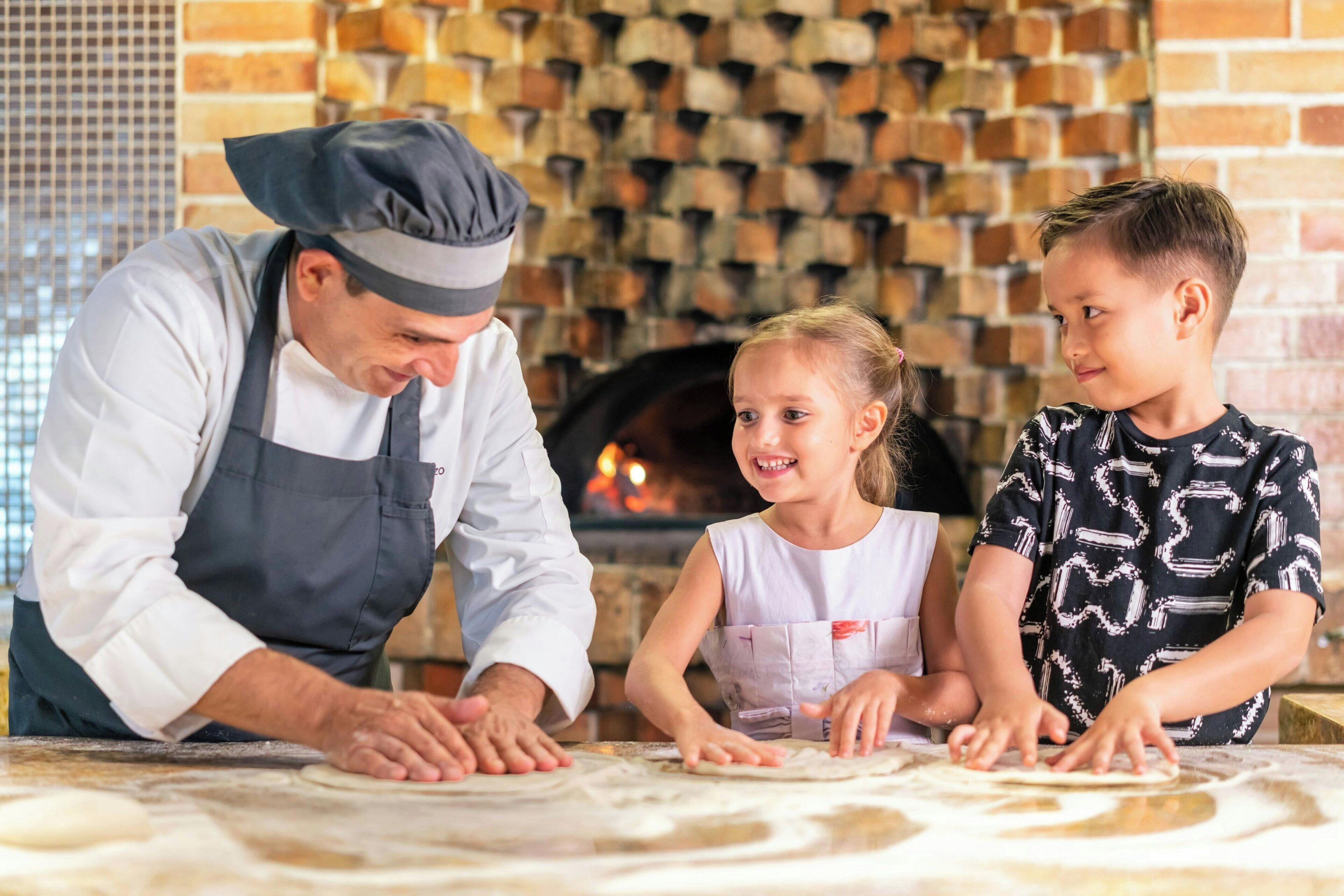 Ein Koch zeigt Kindern, wie man Teig in einem Backofen aus Ziegelsteinen ausrollt.
