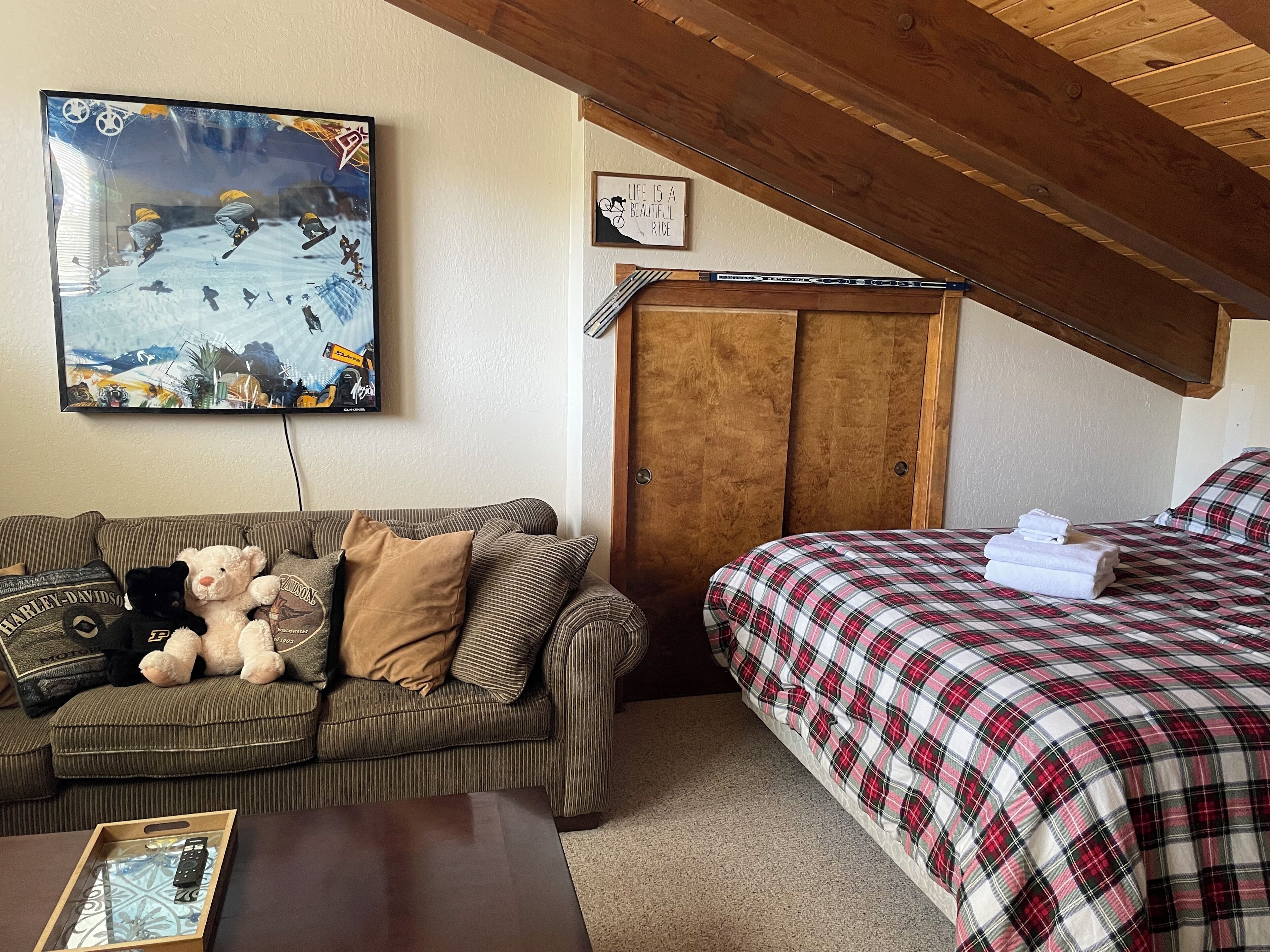 A cozy room with a striped couch, a bed with a plaid bedspread, a wooden wardrobe, a framed quote on the wall, and a painting of a snowy scene.