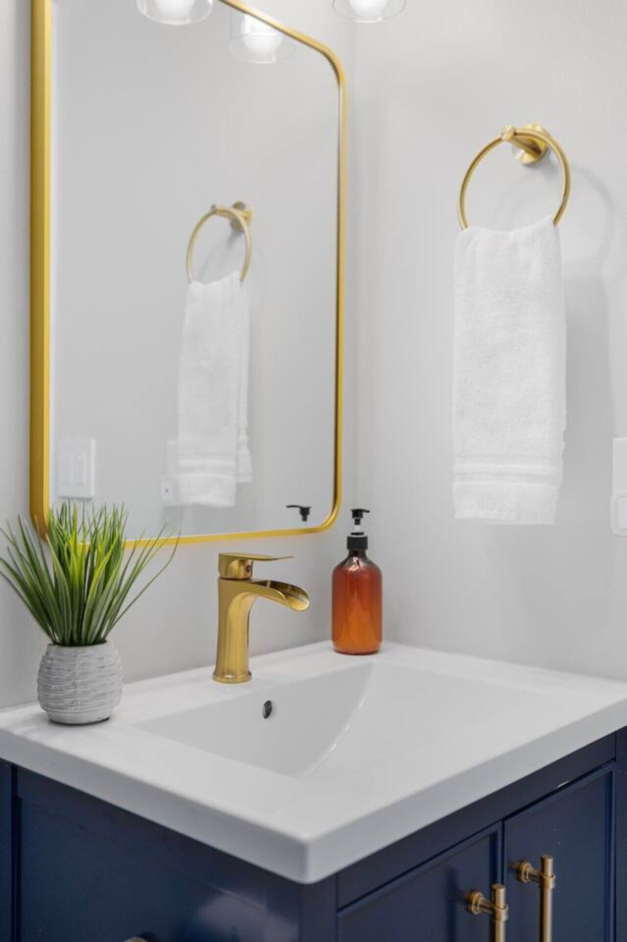 A bathroom with a white sink, gold faucet, and a gold-framed mirror. There is a potted plant on the counter and a bottle of soap.