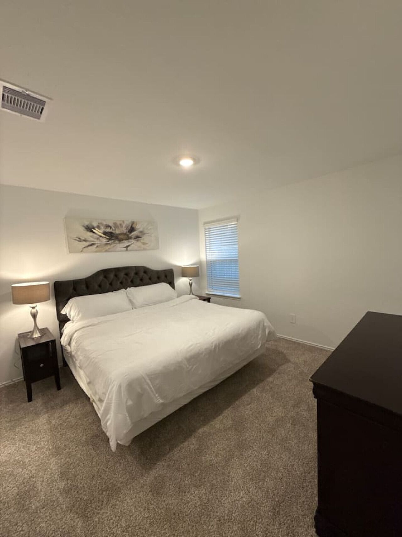 Une chambre à coucher comprenant un lit, une table de chevet, une lampe, un tableau décoratif au mur, une fenêtre avec des stores et une commode sombre.