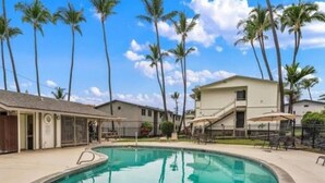 A swimming pool with a surrounding fence, palm trees, and a building with a covered patio area.