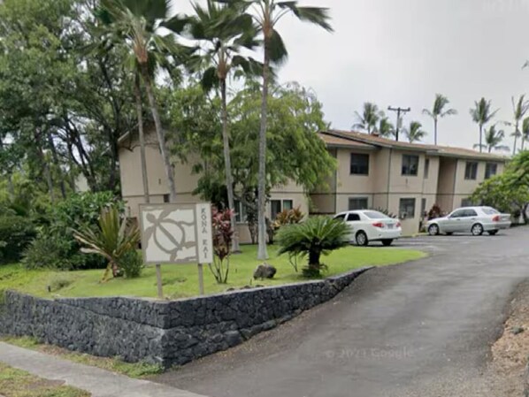 Un edificio con pared de piedra y un letrero, rodeado de palmeras y coches estacionados.