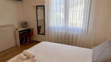 A hotel room with a bed, a desk, a chair, a mirror, and a window with curtains.