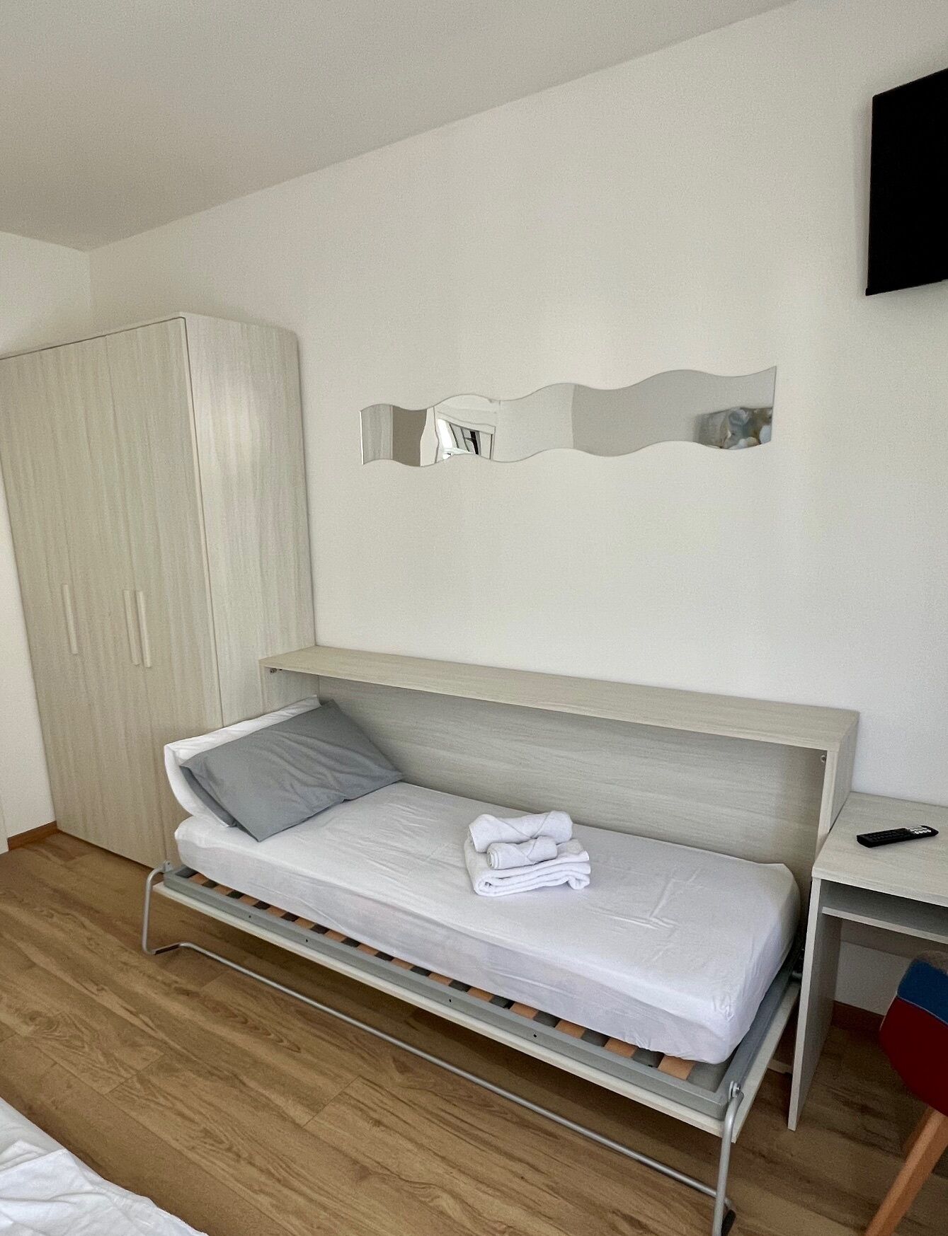 Un lit simple avec un oreiller gris et une serviette pliée, une armoire en bois, une table de chevet avec une télécommande et un miroir fixé au mur.