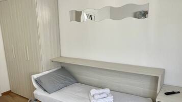 Un lit simple avec un oreiller gris et une serviette pliée, une armoire en bois, une table de chevet avec une télécommande et un miroir fixé au mur.
