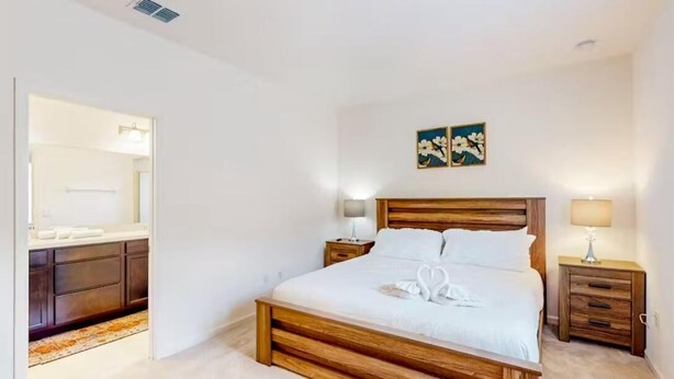 Un dormitorio con una cama de madera, una mesita de noche con lámpara, una obra de arte en la pared y un baño visible a través de una puerta abierta.