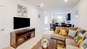 Una sala de estar moderna con un televisor de pantalla plana, un mueble de entretenimiento de madera y un sofá con cojines coloridos. Se ve un comedor con una mesa y sillas al fondo.