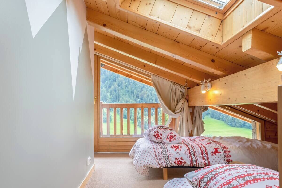 Una camera da letto con due letti, travi in legno al soffitto, un'ampia finestra vista su un paesaggio verdeggiante e un balcone.