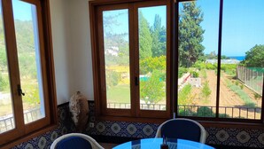 A room with a round glass table and four chairs, large windows with wooden frames, and a view of a garden and coastline.