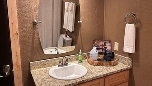 A bathroom with a granite countertop, a sink, a mirror, a towel rack, and a basket with toiletries. - The Sandtown 3 bedroom Cabin (Vian)