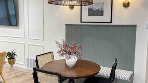 Un comedor con una mesa redonda, dos sillas, una lámpara colgante de fibra, una lámpara de pared y un cuadro enmarcado.