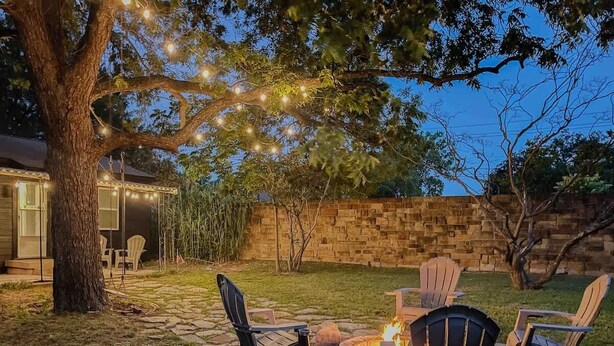 Un área de estar al aire libre con sillas Adirondack y un hogar para fuego, rodeada por un sendero de piedra y una pared de ladrillo.