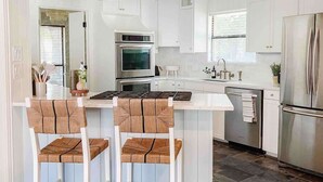 A modern kitchen with white cabinetry, a stainless steel range hood, a built-in oven, and a large refrigerator. There are two woven chairs at the island. - Luxe 4BR with Heated Pool and Hot Tub - Walk to Lake (Grapevine)