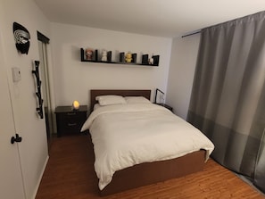Un lit bien fait, avec une literie blanche, une table de chevet avec une lampe, une étagère ornée d’objets décoratifs et une porte avec une poignée.