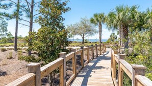 A wooden boardwalk leading to a beach with palm trees and pine trees in the background. - Tropical Treat | Walk to Beach, Pool, & Resort (Port St. Joe)