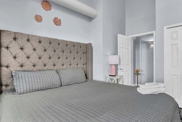 A bedroom with a tufted headboard, a grey quilted bedspread, and a small bedside table with a pink lamp.