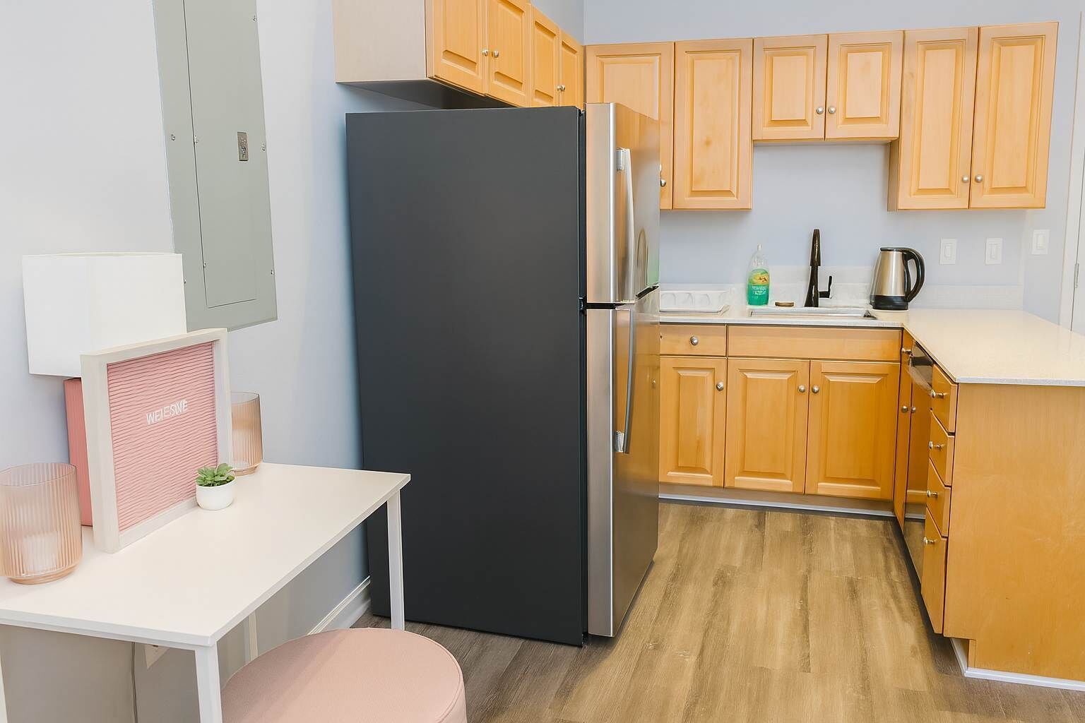 Une cuisine compacte avec des meubles en bois, un réfrigérateur noir, un plan de travail blanc et une petite table accompagnée d’une chaise rose.