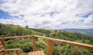 Una terraza de madera con vistas a una ladera verde y exuberante y montañas distantes bajo un cielo parcialmente nublado.