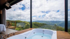 Una habitación con un ventanal grande, una terraza de madera, un jacuzzi y vistas a un paisaje montañoso.